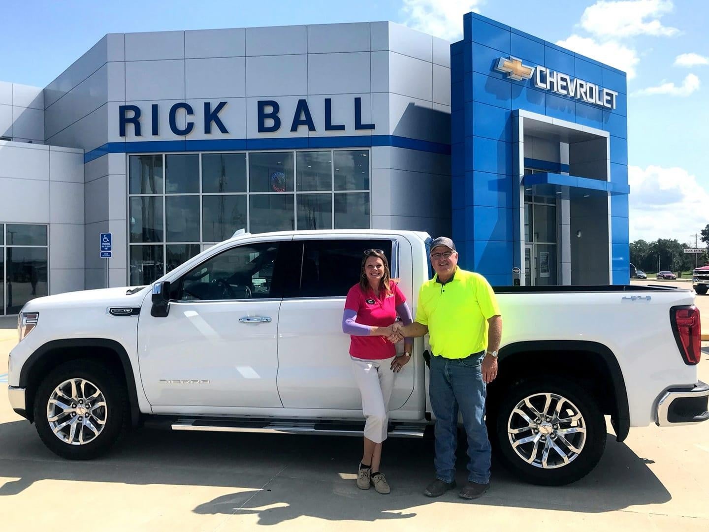 Man and woman shaking hands beside a white pickup truck in front of a blue and white Rick Ball Chevrolet dealership.