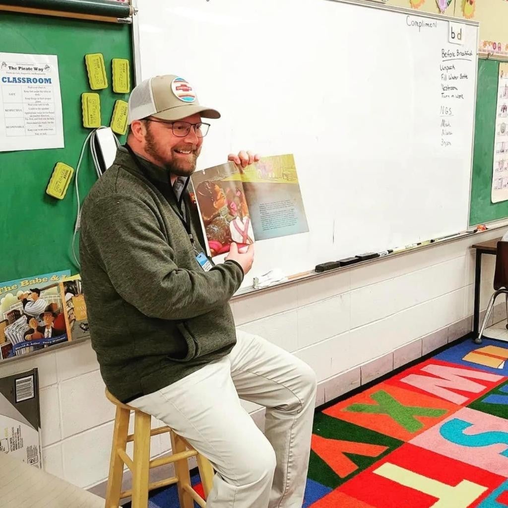 A smiling teacher in a cap and glasses sits on a stool, reading an open children's book to his class.