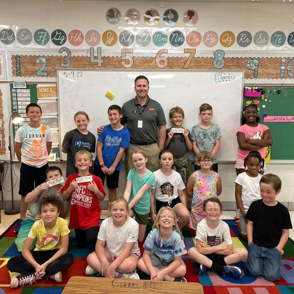 A male teacher and 18 elementary school children pose for a group photo in a colorful classroom.